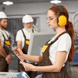 Female engineer in protective ear muffs and glasses holding folder, writing. Engineers wearing in white t shirts, coveralls, helmets. operating computerized plasma laser machine.
