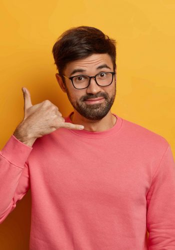 Mobile communication concept. Unshaven man makes call me gesture, talks on imaginary cellular, has friendly positive expression, wears transparent glasses and pink jumper, isolated on yellow wall